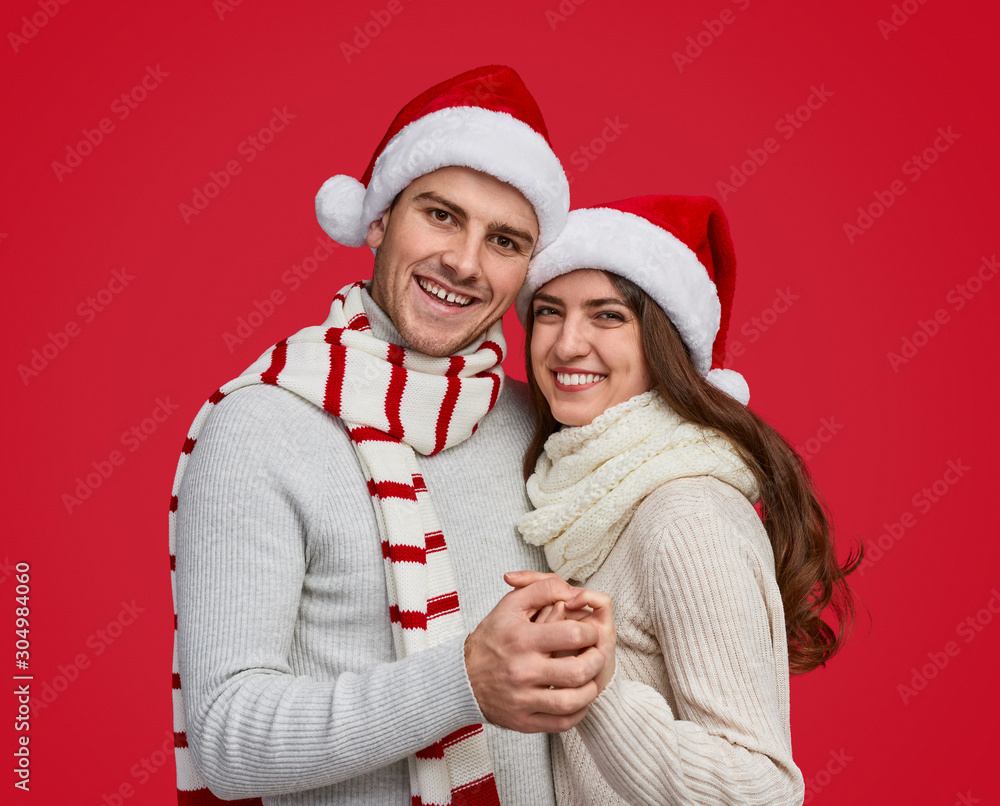 Merry couple in red Christmas hat and scarf hugging and smiling at camera Merry couple in red Christmas hat and scarf hugging and smiling at camera