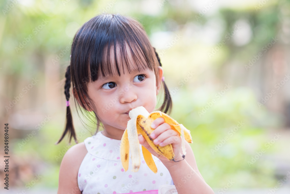 Adorable asian little girl is yummy eating banana, concept of healthy food for kid.