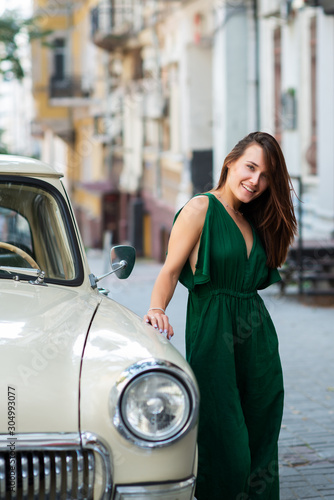Fashion model wearing green overall posing outdoor next to Soviet vintage car. Young beautiful brunette caucasian woman walking summer streets. Beautiful girl, urban portrait.
