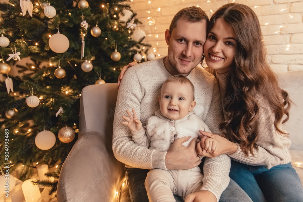 Happy family of mother, father and little baby boy hugging and celebrating new year in front of Christmas tree in decorated interior. Celebrating Christmas. Happy family of mother, father and little baby boy hugging and celebrating new year in front of Christmas tree in decorated interior. Celebrating Christmas.
