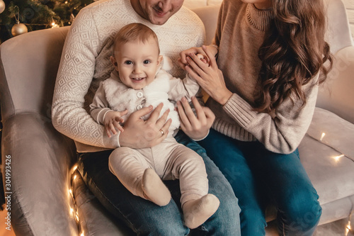 Happy family of mother, father and little baby boy hugging and celebrating new year in front of Christmas tree in decorated interior. Celebrating Christmas.