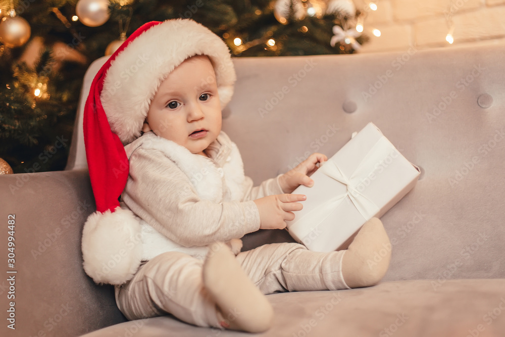 Little baby boy holding gift sitting on sofa in front of Christmas tree .