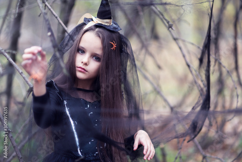 Beautiful little girl with spider in her long brunette hair, dressed in a witch dress and hat walks in fall forest with fear expressions. Halloween horror. Witches in darkness photo