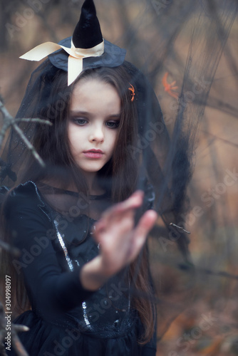 Llittle girl dressed in a witch dress and hat walks in fall forest among the web with spiders. Halloween horror. Witches in darkness photo