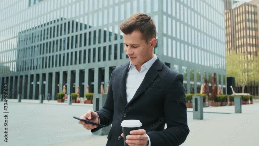 Handsome young Caucasian man in business style tapping and texting on the smartphone while standing in the city with a coffee to-go in hand. Outside. Handsome young Caucasian man in business style tapping and texting on the smartphone while standing in the city with a coffee to-go in hand. Outside.