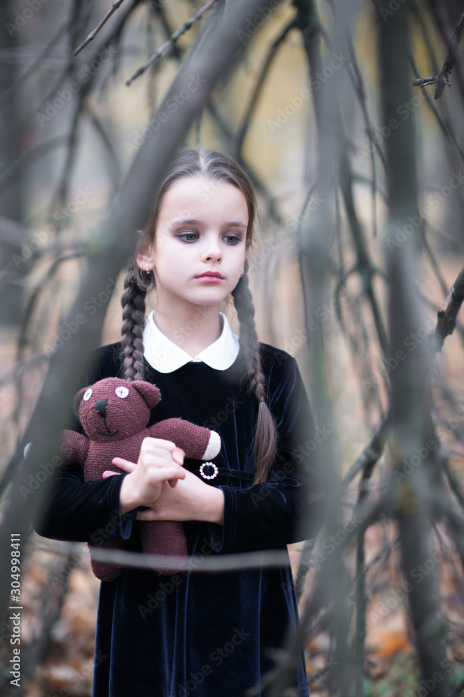 Beautiful little girl with long brunette hair, dressed in black velvet dress walks in fall forest with handmade bear toy. Halloween horror,  ghost or spirit of child in twilight