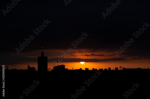 Sun appears from behind the clouds, above the city, roof top view of an old church tower. Summer sunrise or sunset. Dark orange sky color tones. Old town of Badalona, Spain photo