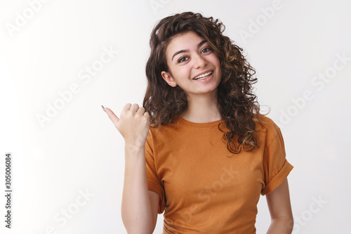 Girl inviting go together. Friendly charming smiling cute armenian woman tilting head pointing left thumb joyfully grinning asking you wanna visit cool place, recommending cafe, standing white wall photo