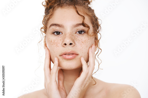 Headshot sensual gentle redhead curly-haired woman freckles standing naked white background touching pure clean skin look mirror applying skincare treatment cosmetology products, white background