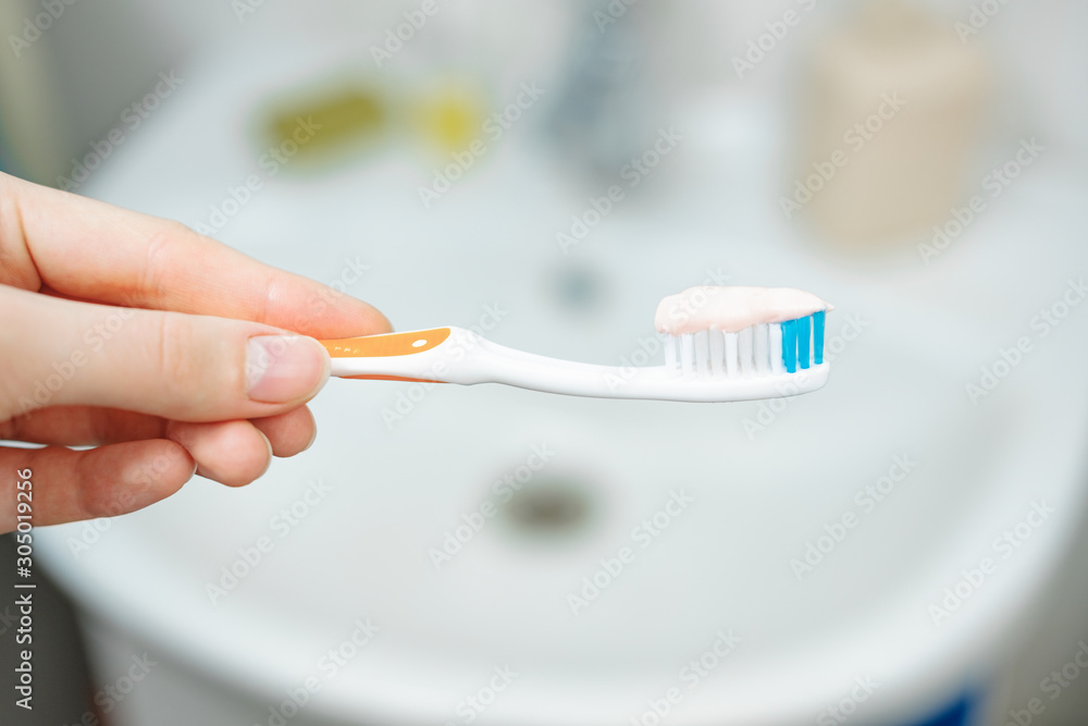 Hand holds a toothbrush with toothpaste on bathroom background, close up view. Dental care and oral hygiene concept.