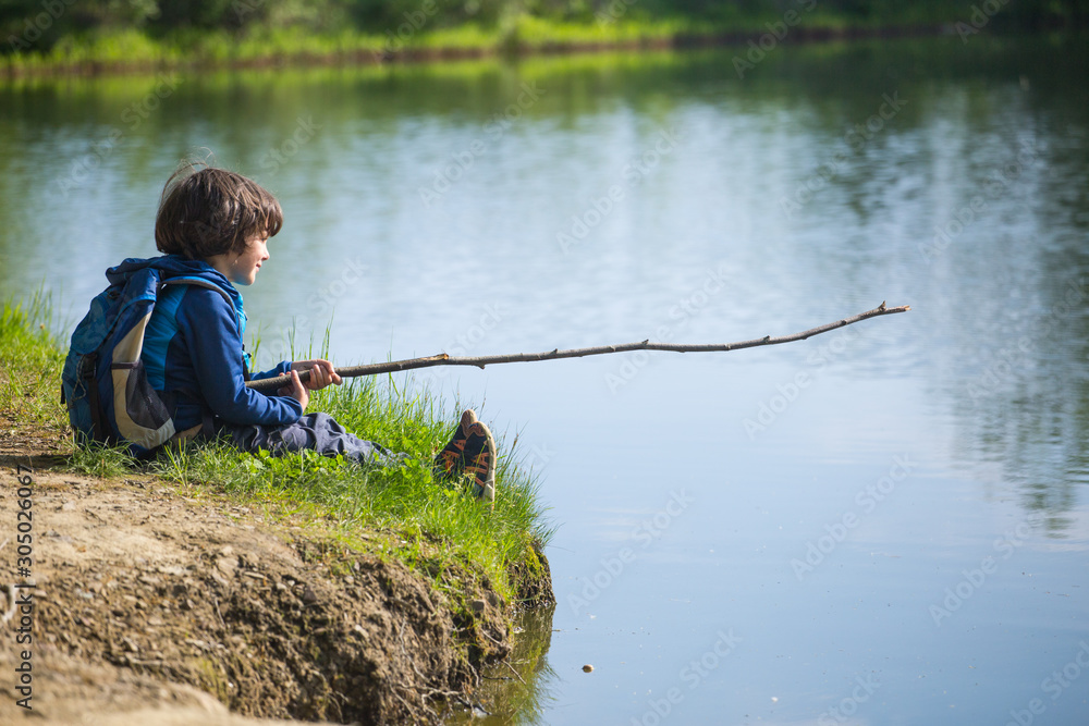 A boy with a backpack sits on the lake with a wooden stick. A boy with a backpack sits on the lake with a wooden stick.