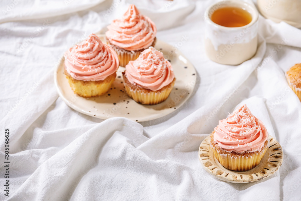 Homemade cupcakes with pink buttercream and coconut flakes served with ceramic teapot, cup of tea on white folded tablecloth. Homemade cupcakes with pink buttercream and coconut flakes served with ceramic teapot, cup of tea on white folded tablecloth.