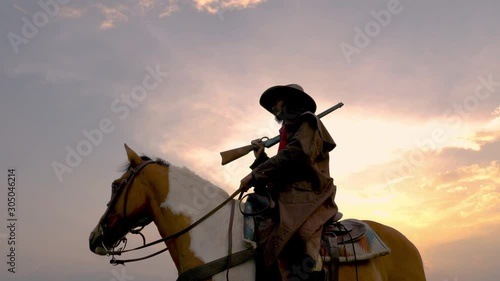Cowboy riding across grassland with lake behind, Cowboy ranger or hunter with gun walking in forest early afternoon. Cowboy on horse riding and holding a gun in silhouette Scene.Panning around cowboy.
