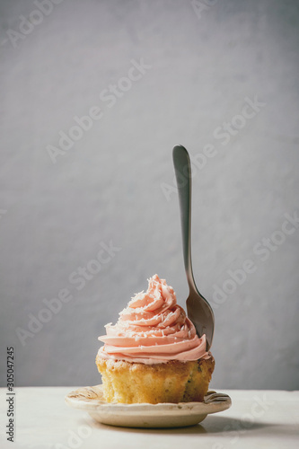 Homemade cupcake with pink buttercream and coconut flakes served on ceramic plate with dessert fork on white marble table. Copy space.