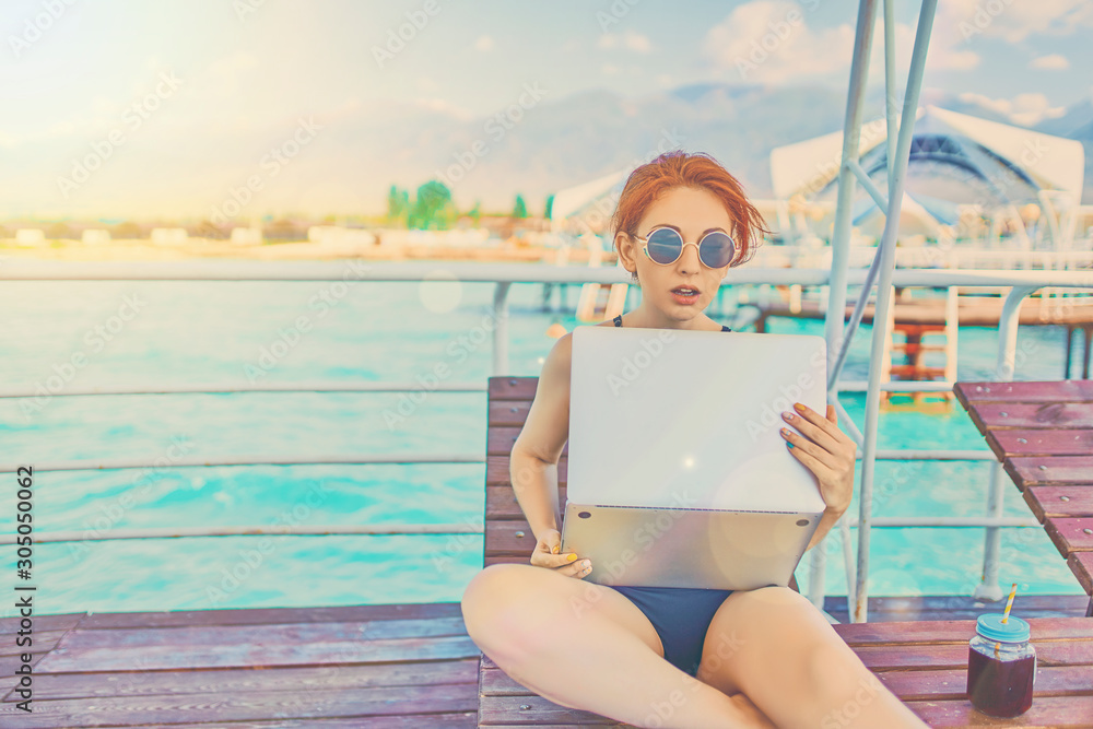 Redhead girl working with a laptop on the sea. Female freelancer on the beach.