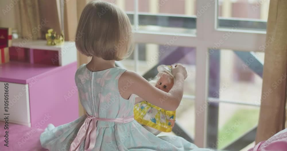 Top back view of little Caucasian blond girl playing with doll as sitting at pink bed. Child in beautiful blue dress with pink butterflies hugging the toy and fixing its hat. Childhood, leisure. Top back view of little Caucasian blond girl playing with doll as sitting at pink bed. Child in beautiful blue dress with pink butterflies hugging the toy and fixing its hat. Childhood, leisure.