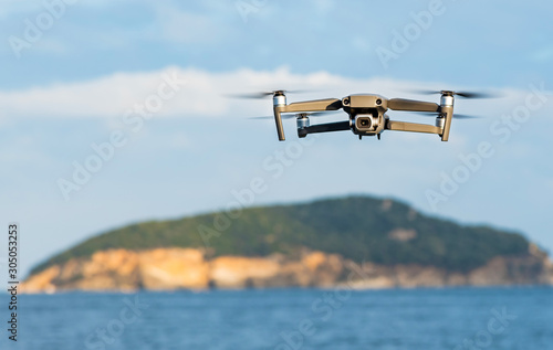 Drone with camera hovering in the mid air outdoors, island and sea on background.