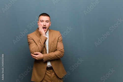 young handsome man open-mouthed in shock and disbelief, with hand on cheek and arm crossed, feeling stupefied and amazed against flat wall photo