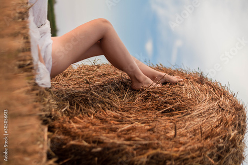 Legs of a young gentle girl against a background of a round bundle of hay in an open field in the sunset light