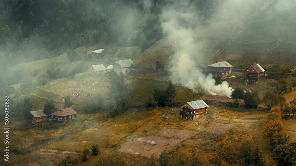 Fototapeta premium Villagers burn foliage and grass in their gardens in the fall, smoke and fog rises above the village, hills and mountains.