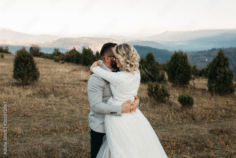 Beautiful couple having a romantic moment on their weeding day, in mountains at sunset. Bride is in a white wedding dress with a bouquet of sunflowers in hand, groom in a suit. Happy hugging couple.