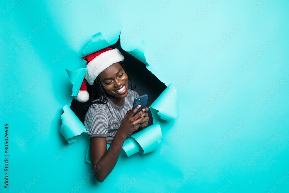 Obraz premium Young african woman wear in santa hat use phone in hands standing in green torn paper hole background