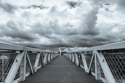 Fotografía en blanco y negro de vista en primera persona caminando por pasarela peatonal metalica con fondo de nubes dramaticas