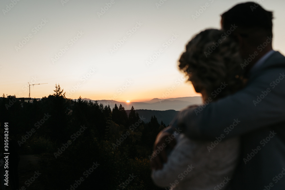 Beautiful couple having a romantic moment on their weeding day, in mountains at sunset.Bride is in a white wedding dress with a bouquet of sunflowers in hand, groom in a suit. Watching sunset together