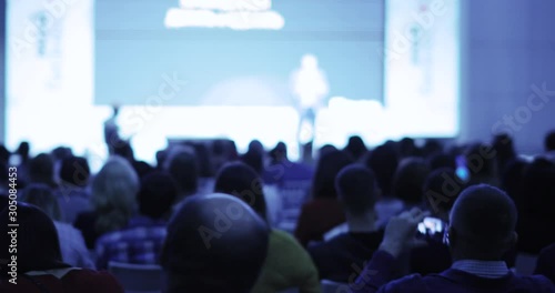 Unrecognizable businessman on stage talking and showing presentation on screen to large audience sitting back to camera, blur background. Training, conference, public speech concept.