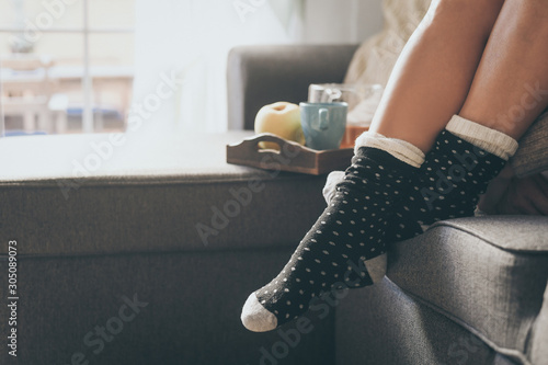 Closeup view of female legs and feet with warm socks in a winter morning. Woman sitting on a couch at home having natural breakfast with tea. Girl resting on the sofa in cozy soft comfortable sweater.