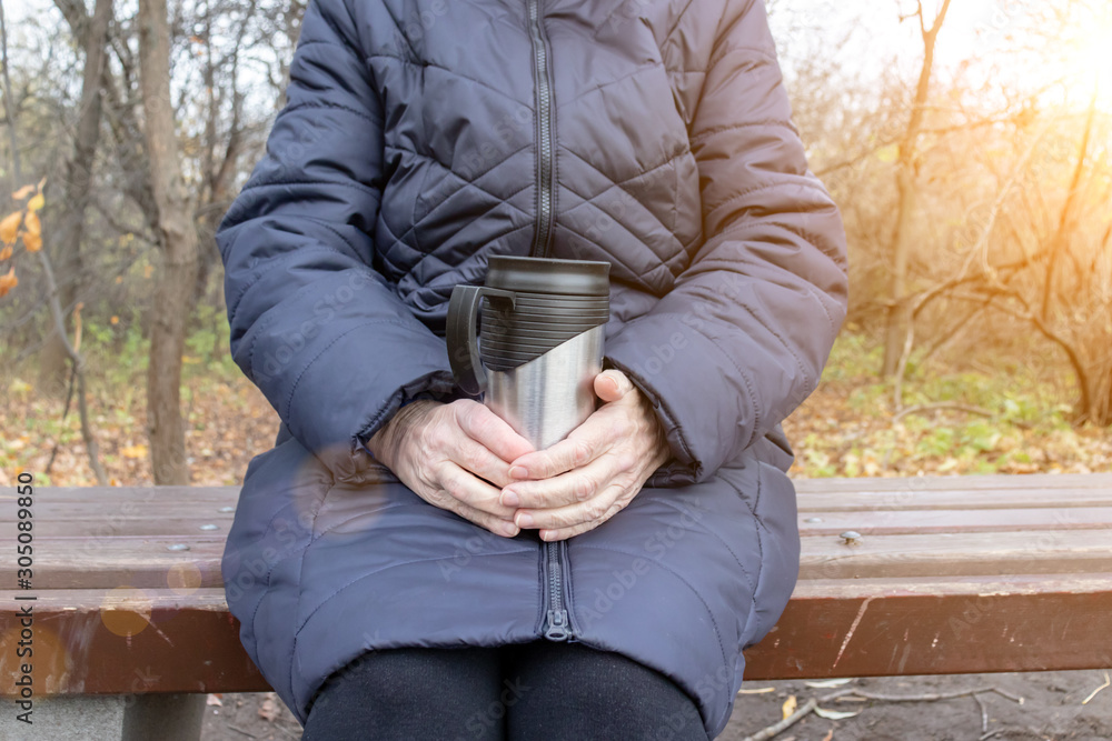 An old senior woman sitting on a bench in late autumn park and holding thermo cup in hands, hot drinks outdoor concept An old senior woman sitting on a bench in late autumn park and holding thermo cup in hands, hot drinks outdoor concept