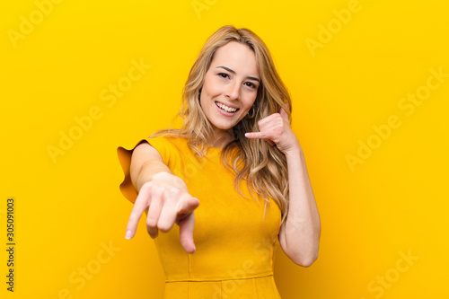 young pretty blonde woman smiling cheerfully and pointing to camera while making a call you later gesture, talking on phone against flat color wall photo