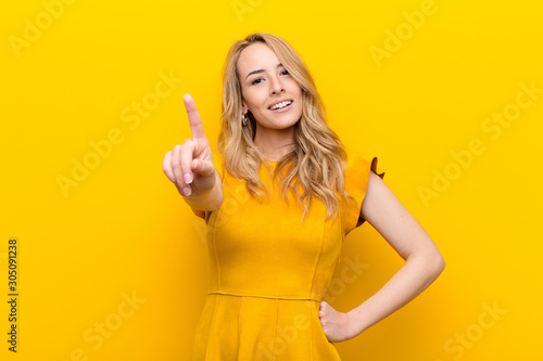 young pretty blonde woman smiling and looking friendly, showing number one or first with hand forward, counting down against flat color wall