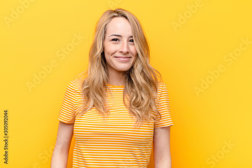 young pretty blonde woman smiling positively and confidently, looking satisfied, friendly and happy against flat color wall