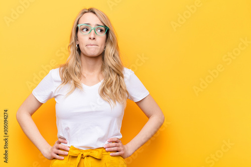 young pretty blonde woman doubting or thinking, biting lip and feeling insecure and nervous, looking to copy space on the side against flat color wall photo