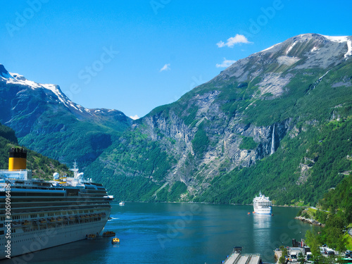 Popular Geiranger Fjord and surrounding mountains with snow, waterfalls, moored cruise ships and tender boats carrying tourists from ship to shore on a beautiful day with blue sky.