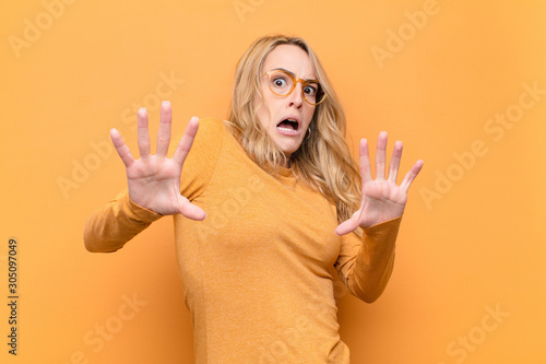 young pretty blonde woman feeling terrified, backing off and screaming in horror and panic, reacting to a nightmare against flat color wall photo