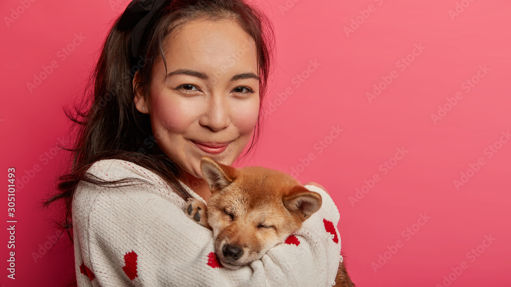 Close up shot of pretty satisfied Asian woman takes care of pet, embraces with love shiba inu dog, gets animal as present, stands against pink background with empty space. Enjoying time together Close up shot of pretty satisfied Asian woman takes care of pet, embraces with love shiba inu dog, gets animal as present, stands against pink background with empty space. Enjoying time together