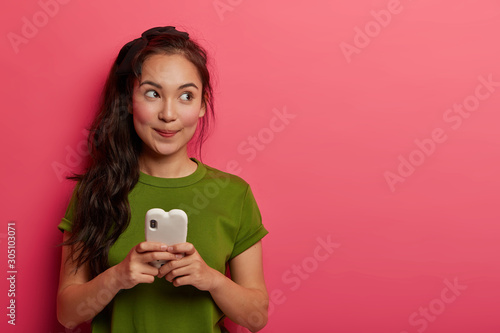 Studio shot of positive thoughtful korean girl uses mobile phone for chatting online, looks away, wears casual clothing, downloads music for playlist, plays app, isolated over pink background