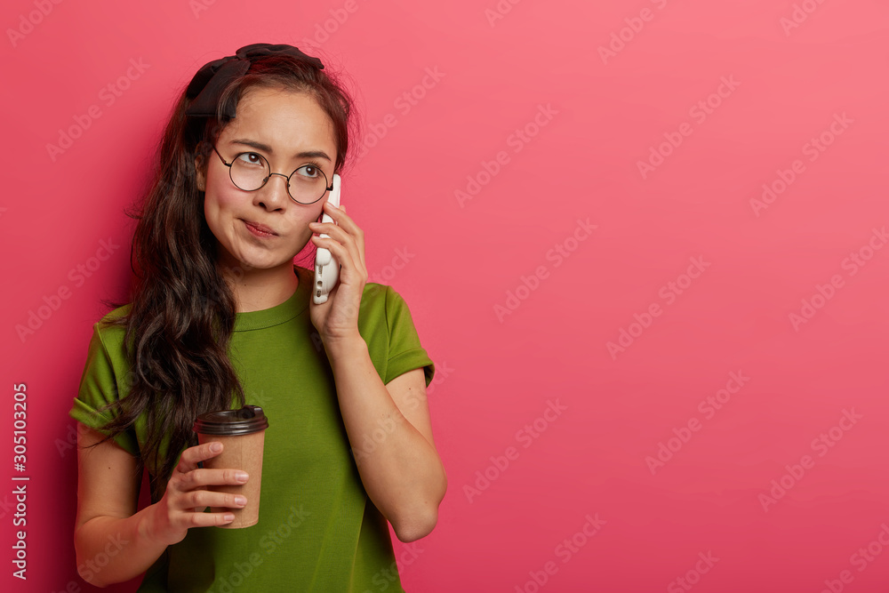 Millennial brunette female makes phone call, tired of waiting someone answers, keeps cellular near ear, drinks takeaway coffee, dressed in casual green t shirt, poses indoor. Boring conversation Millennial brunette female makes phone call, tired of waiting someone answers, keeps cellular near ear, drinks takeaway coffee, dressed in casual green t shirt, poses indoor. Boring conversation