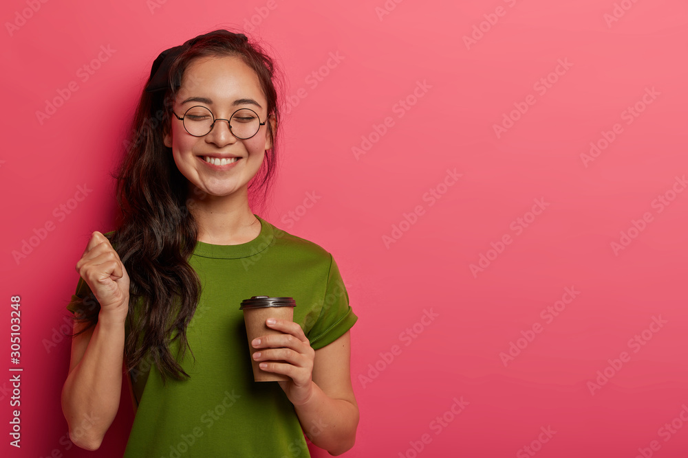 Studio shot of joyful student raises fist, rejoices triumph and successfully finished task, has coffee break after long hours of work, smiles positively, wears eyeglasses, t shirt, poses with beverage Studio shot of joyful student raises fist, rejoices triumph and successfully finished task, has coffee break after long hours of work, smiles positively, wears eyeglasses, t shirt, poses with beverage
