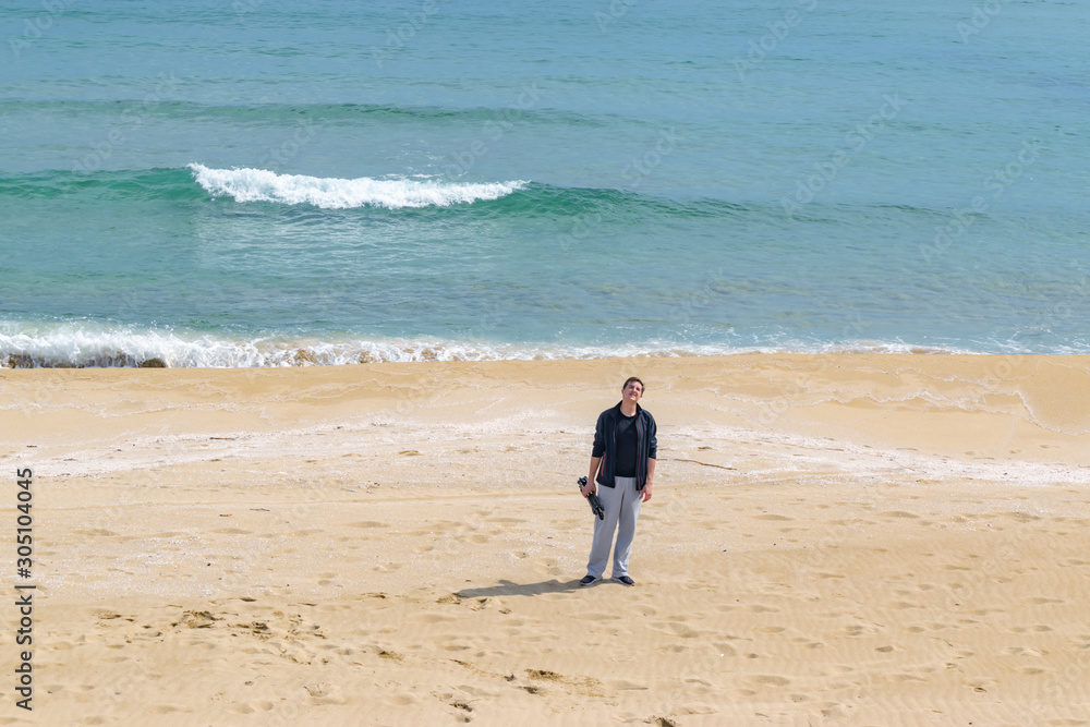 Young man photographer with tripod in hand standing in the middle of sandy beach with sea and waves on background. Travel photographer concept. Copy space, horizontal image. Young man photographer with tripod in hand standing in the middle of sandy beach with sea and waves on background. Travel photographer concept. Copy space, horizontal image.