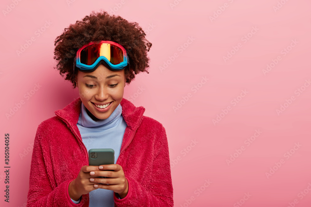 Active female skier or snowboarder looks gladfully at mobile phone, gets nice notification, shares photos after unforgettable journey or holiday, wears ski goggles on head, isolated on pink wall