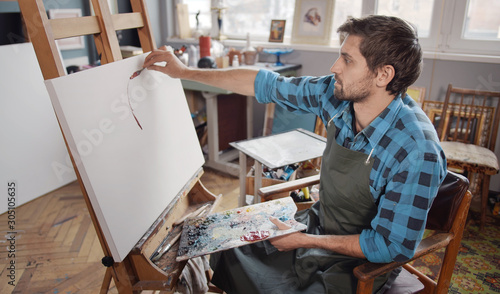 Inspired male artist in blue checked shirt making a first stroke of his new painting in a light loft studio