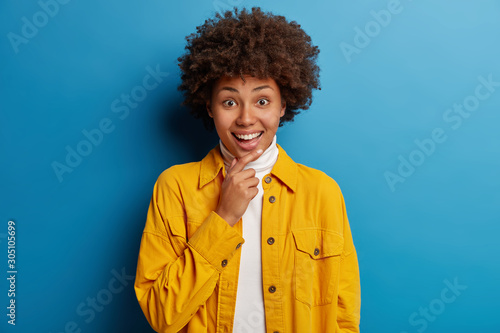 Cute smiling Afro American, holds chin, looks with glad surprised expression at camera, hears pleasant news, wears bright yellow jacket, isolated over blue background. Human emotions and feelings