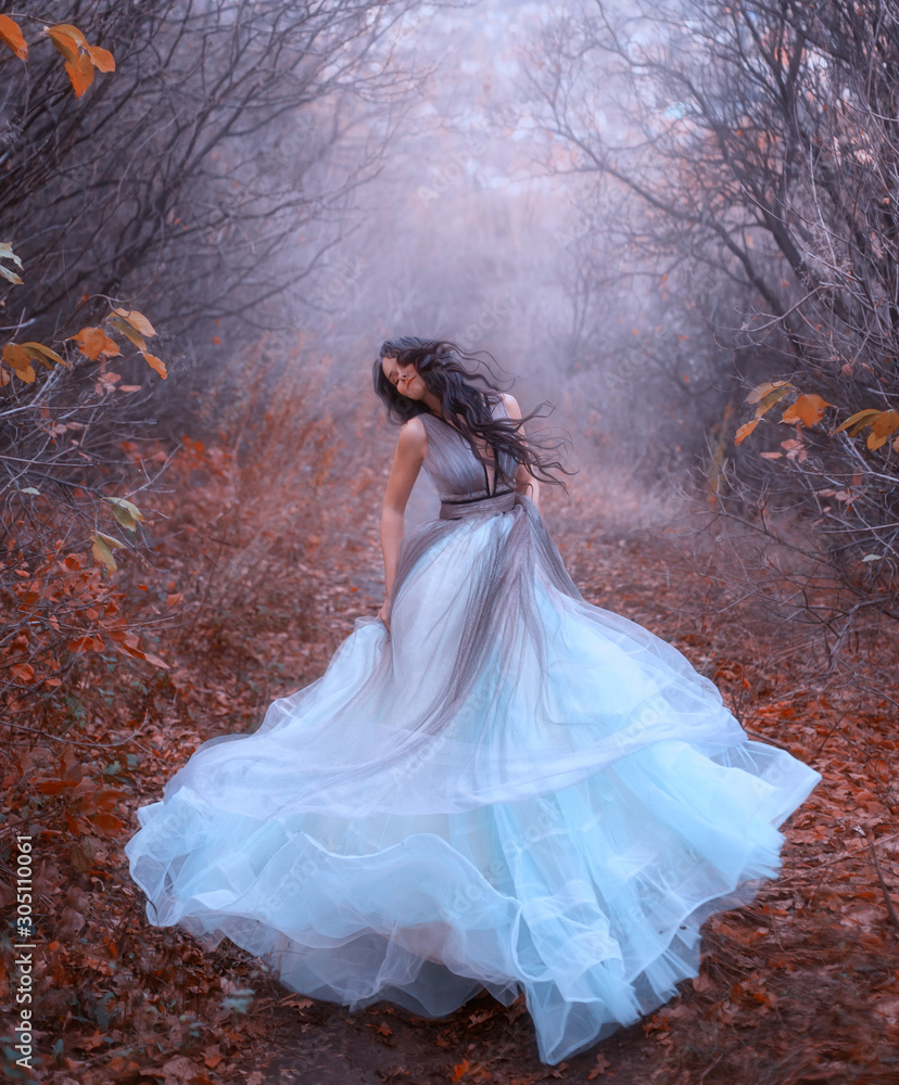 Fototapeta premium Piękna fantazja kobieta kręci się w tańcu. Luksusowa niebieska sukienka w stylu art. Czarne, długie włosy fruwają na wietrze. Księżniczka spaceruje po jesiennym lesie. Tło mgła nagie drzewa opadłe liście pomarańczy