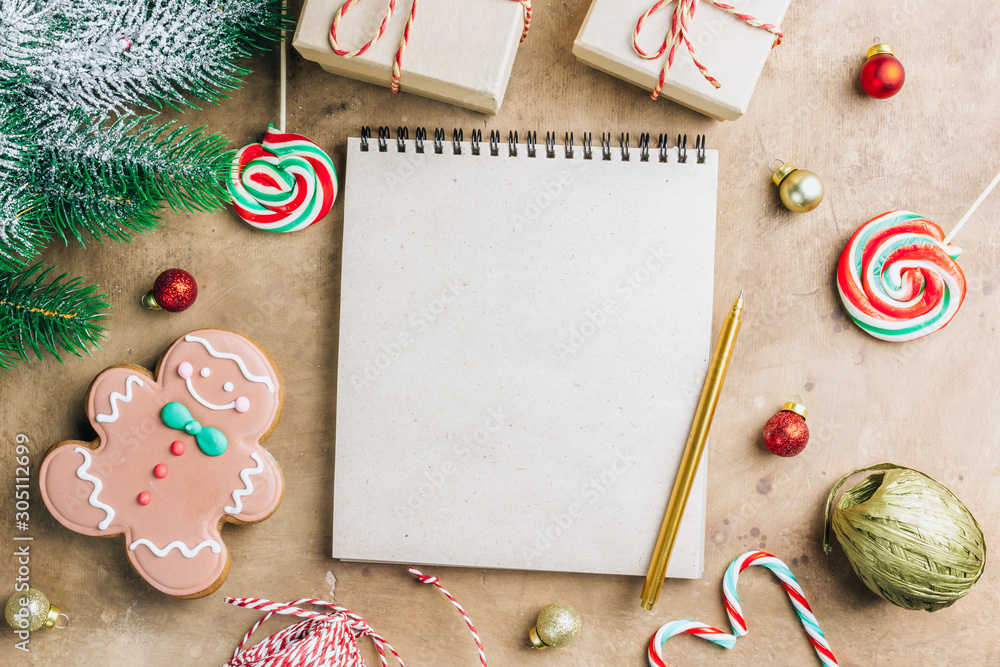 Notebook in the center of cute Christmas decoration: gift box, christmas balls, gingerbread man, fir tree branch, cande cane. Flat lay. Top view Notebook in the center of cute Christmas decoration: gift box, christmas balls, gingerbread man, fir tree branch, cande cane. Flat lay. Top view
