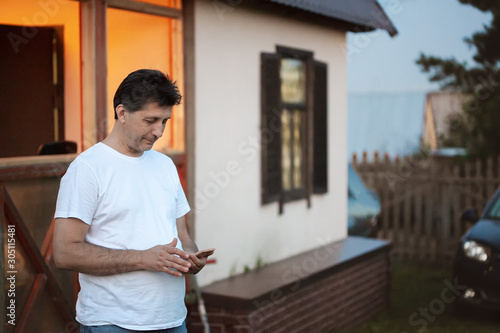 family holidays in the country. white middle-aged man stands near the house looking at the phone. summer evening, in the background a car.