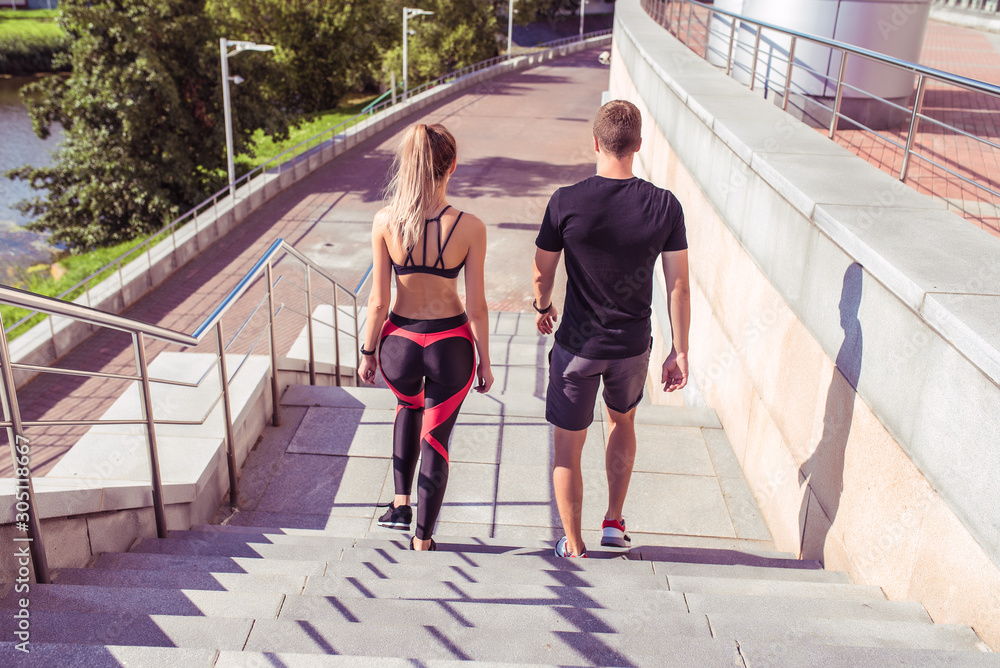 Athletic and young fitness couple descends stairs, rest after jogging and fitness training in the summer in the city, view from the back, sportswear. Background stairs trees and lake with a river.