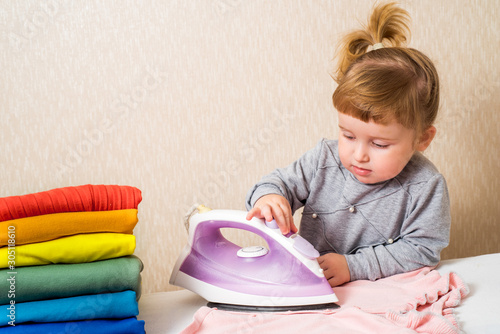baby girl ironing baby clothes. a stack of bright clothes like a rainbow. Housewife helps mom